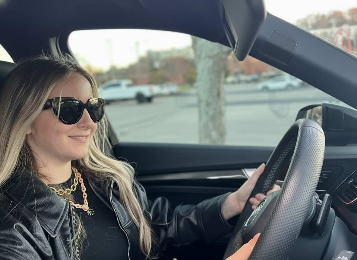 Woman driving a car with sunglasses on, parked in a lot.